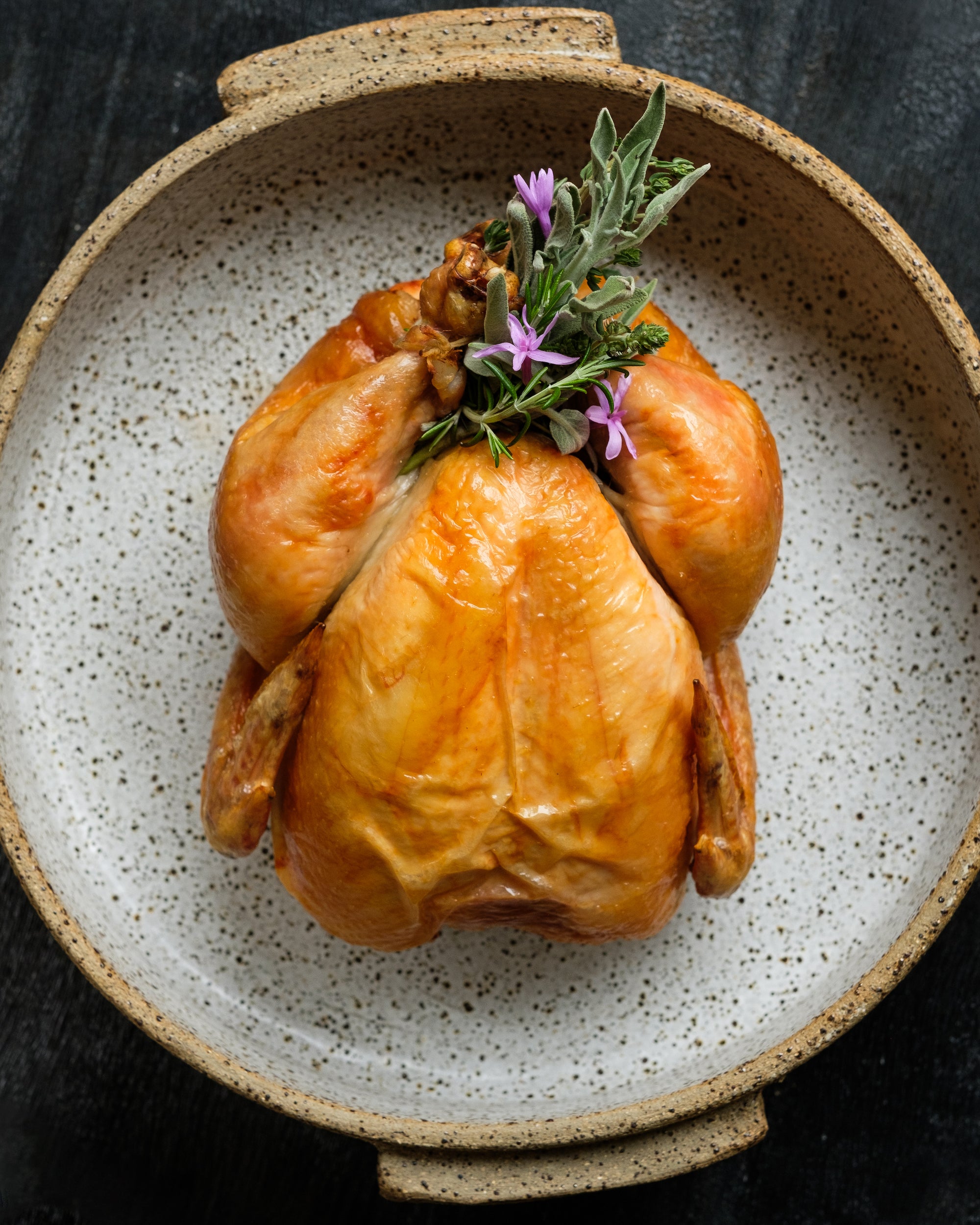 Whole roasted chicken garnished with fresh herbs in a speckled ceramic dish, representing high-quality grass-fed meats for a nutritious and flavorful meal.
