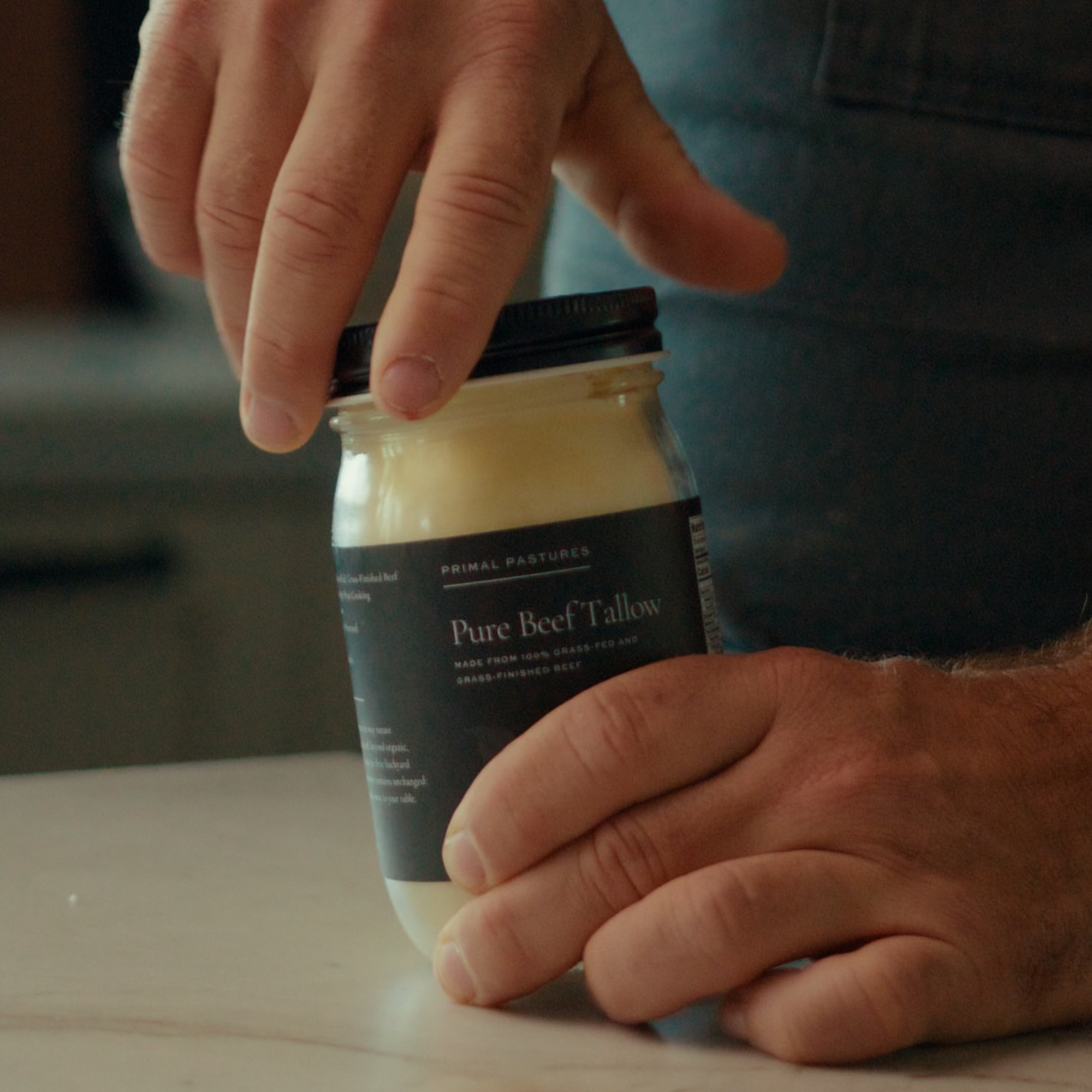 Hands holding a jar of Primal Pastures Pure Beef Tallow on a smooth surface.