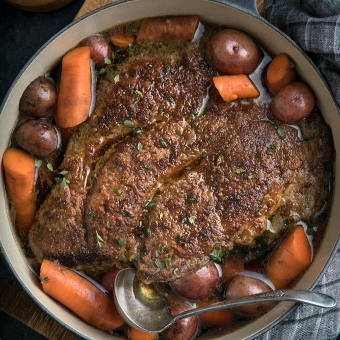 A cooked beef chuck roast in a roasting pan with carrots and potatoes, garnished with herbs.