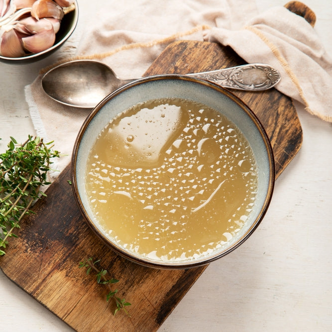 A bowl of beef bone broth on a wooden board with a spoon and parsley on the side, indicating a nutritious and tasty soup base.