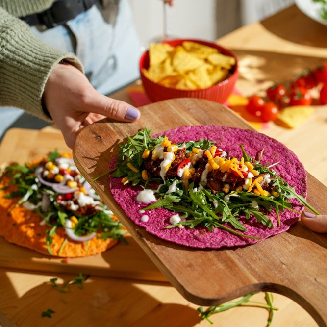 A person holding a tortilla with beef chorizo, greens, and tomato salsa on a wooden board, with various ingredients and dishes in the background.
