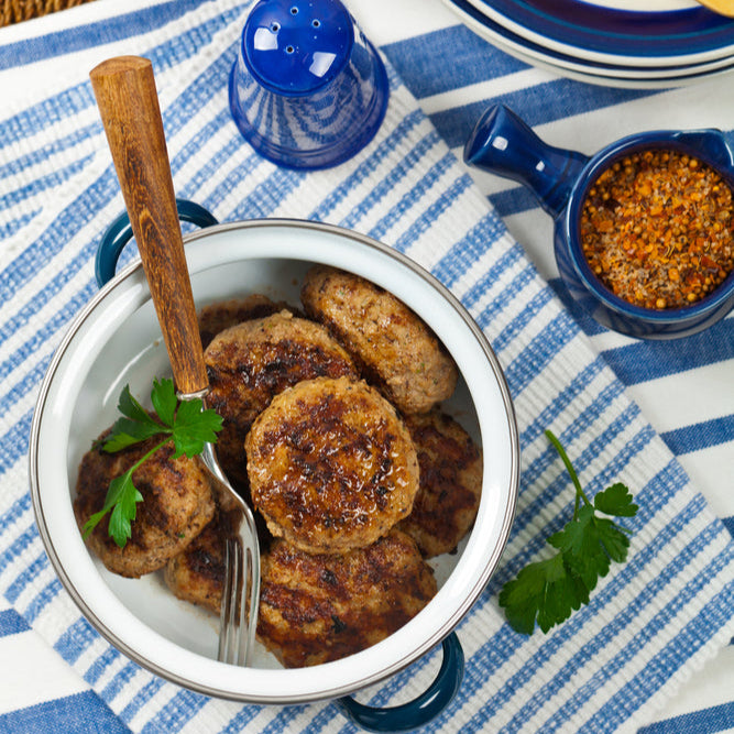 A bowl of cooked ground pork sausage patties on a blue and white striped kitchen towel, with seasonings visible on the patties and a fork resting inside the bowl.