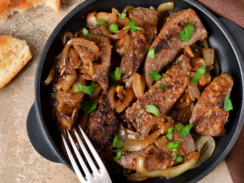 A skillet filled with tender, pan-fried grass-fed beef liver, garnished with sautéed onions and fresh parsley.
