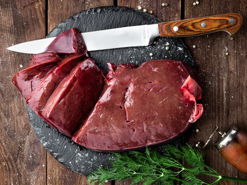 Freshly sliced grass-fed beef liver on a black slate board, accompanied by a knife and garnished with dill.