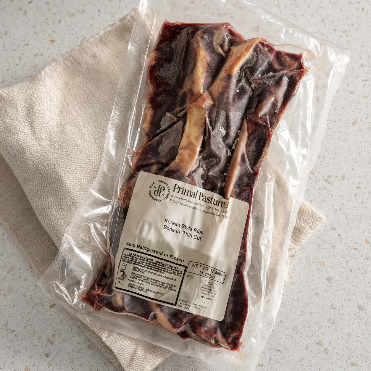 A vacuum-sealed package of Primal Pasture Korean Bone-in Spare Rib Thick Cut raw beef, resting on a beige cloth over a speckled white countertop.