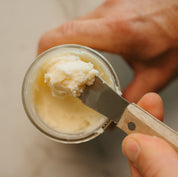Hand scooping beef tallow from a glass jar with a butter knife.