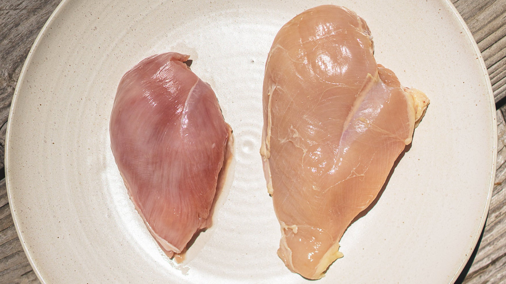 Two pieces of raw chicken breast on a white plate with a wooden surface underneath.