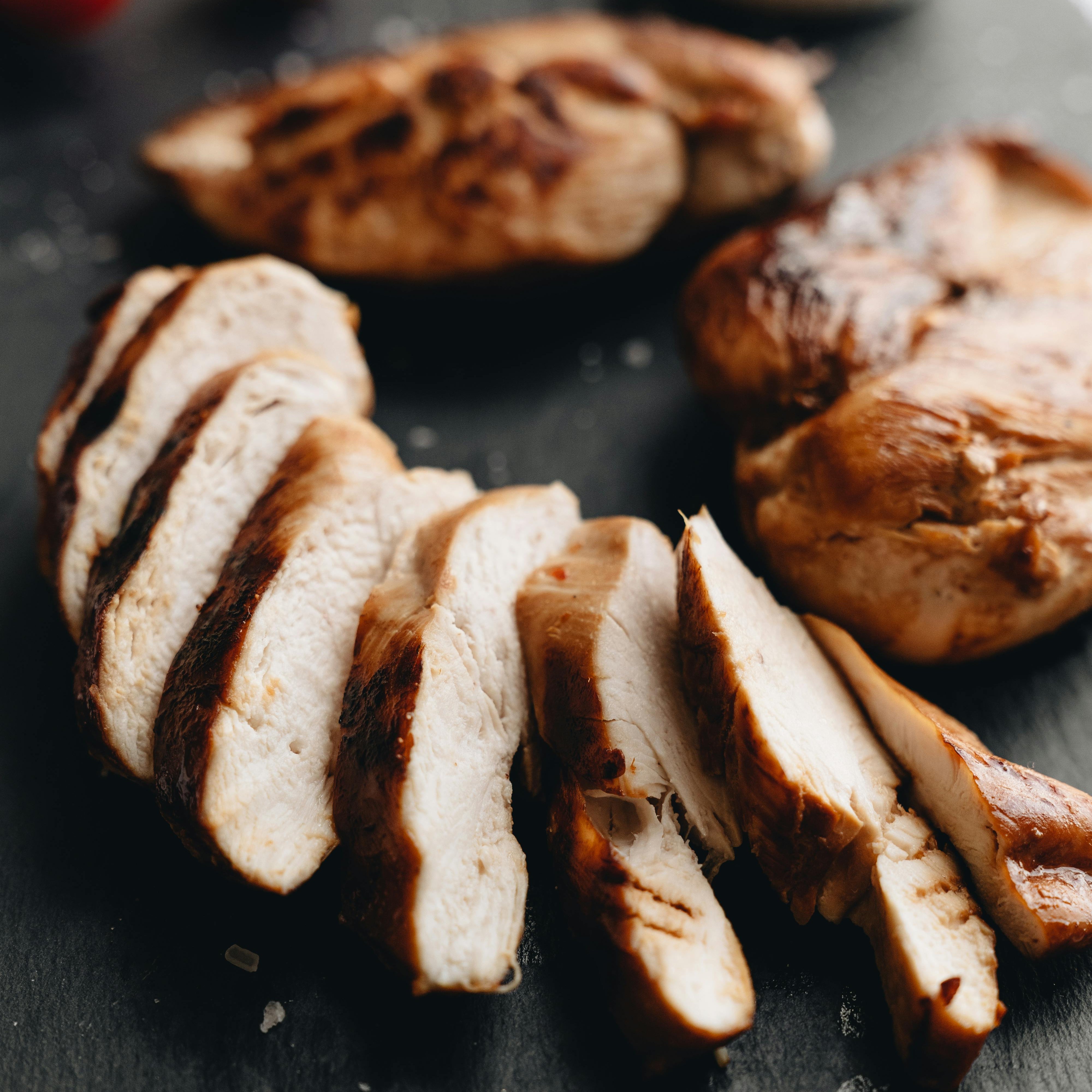 Sliced grilled chicken breast on a dark surface with cherry tomatoes and a small bowl of sauce in the background.