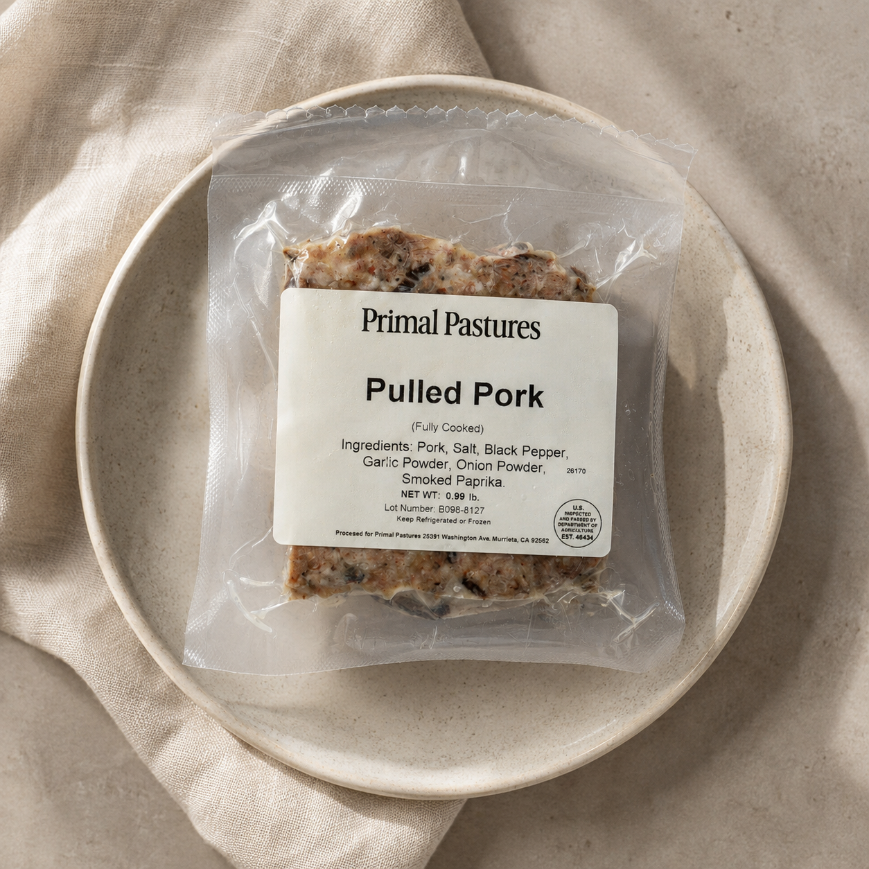A vacuum-sealed package of Primal Pastures Pulled Pork resting on a light beige linen cloth inside a speckled ceramic plate. The package features a white label detailing the ingredients and product information.
