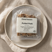 A vacuum-sealed package of Primal Pastures Pulled Pork resting on a light beige linen cloth inside a speckled ceramic plate. The package features a white label detailing the ingredients and product information.