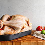 A raw, seasoned whole turkey in a black roasting pan sits on a wooden cutting board, with apples, oranges, and limes on a beige cloth and wooden board to the right against a textured gray wall.