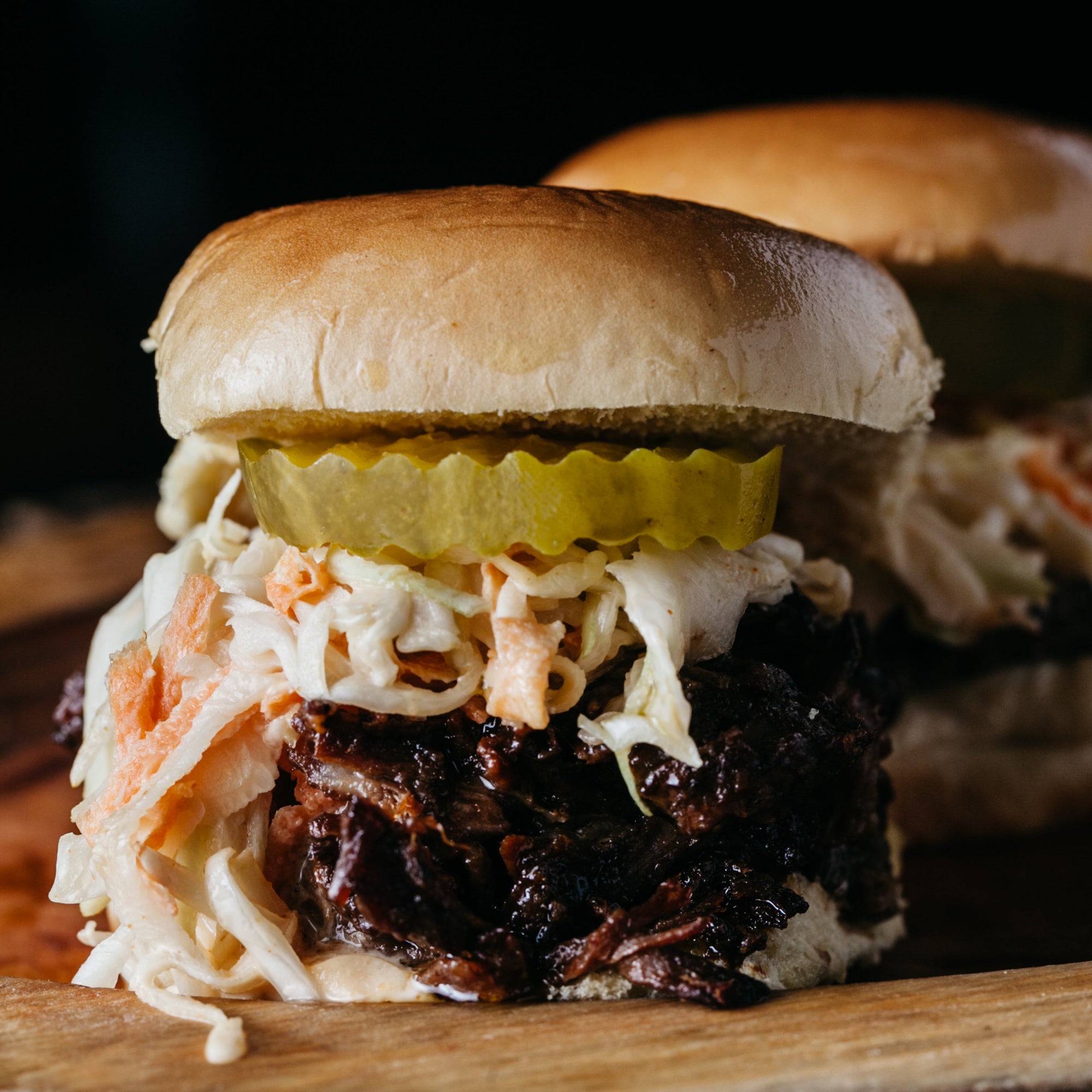 Close-up of a slider with coleslaw and a pickle on a wooden surface.