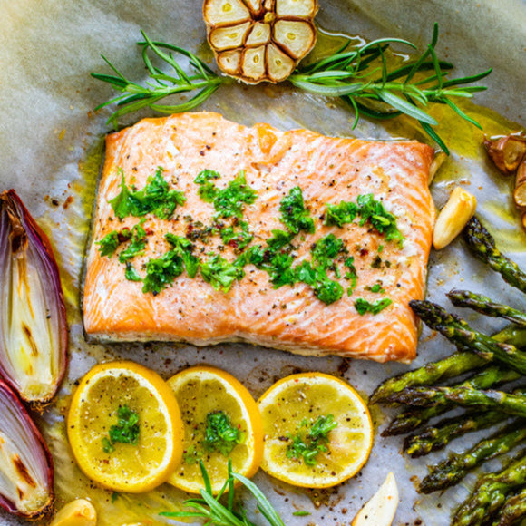 A prepared wild steelhead trout meal on a baking sheet, including filets of trout garnished with herbs, roasted vegetables, and lemon slices.