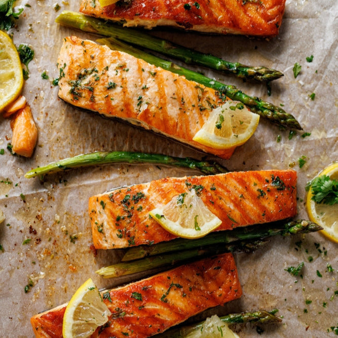 Cooked salmon filets on a baking sheet with lemon slices and green herbs, possibly dill, scattered around. The salmon is pink and appears to be seasoned.